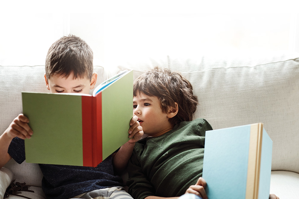 Gifts from the Heart - Two boys sitting on a couch reading books. Younger brother looking on from older sibling