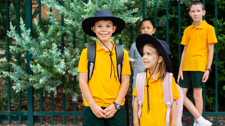 Primary school children in yellow uniforms wearing hats and backpacks, smiling outside a school