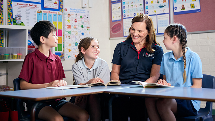 A teacher sits at a table with three primary school students, smiling and reading together in a classroom.