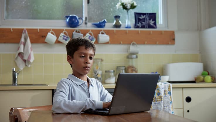 young boy sits at laptop doing homework in kitchen