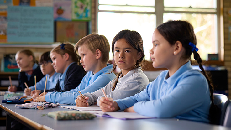 a school girl in a classroom sitting next to her classmates. She is looking directly at the camera while her classmates look ahead