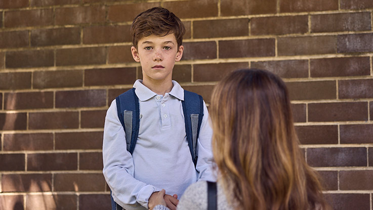 a school boy looking at the camera directly while his mum is kneeling down in front of him with her back turned