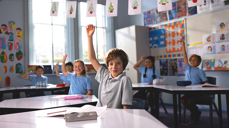 Primary school students sitting at desks in a classroom, with one student raising a hand to answer a question.