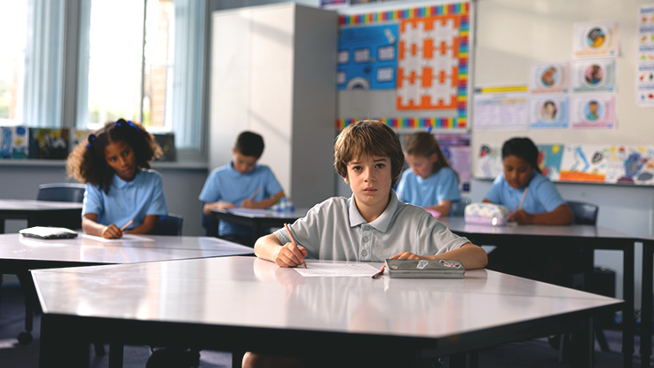 Primary school students sitting at desks working on assignments in a classroom with educational posters on the walls.