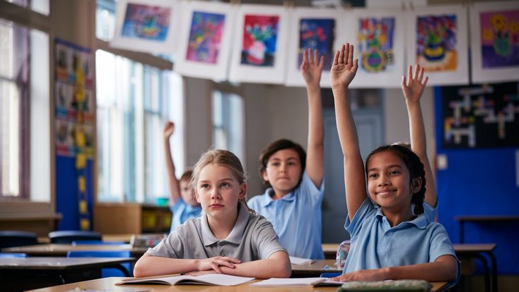 Students seated at desks in a classroom, with several students raising their hands.