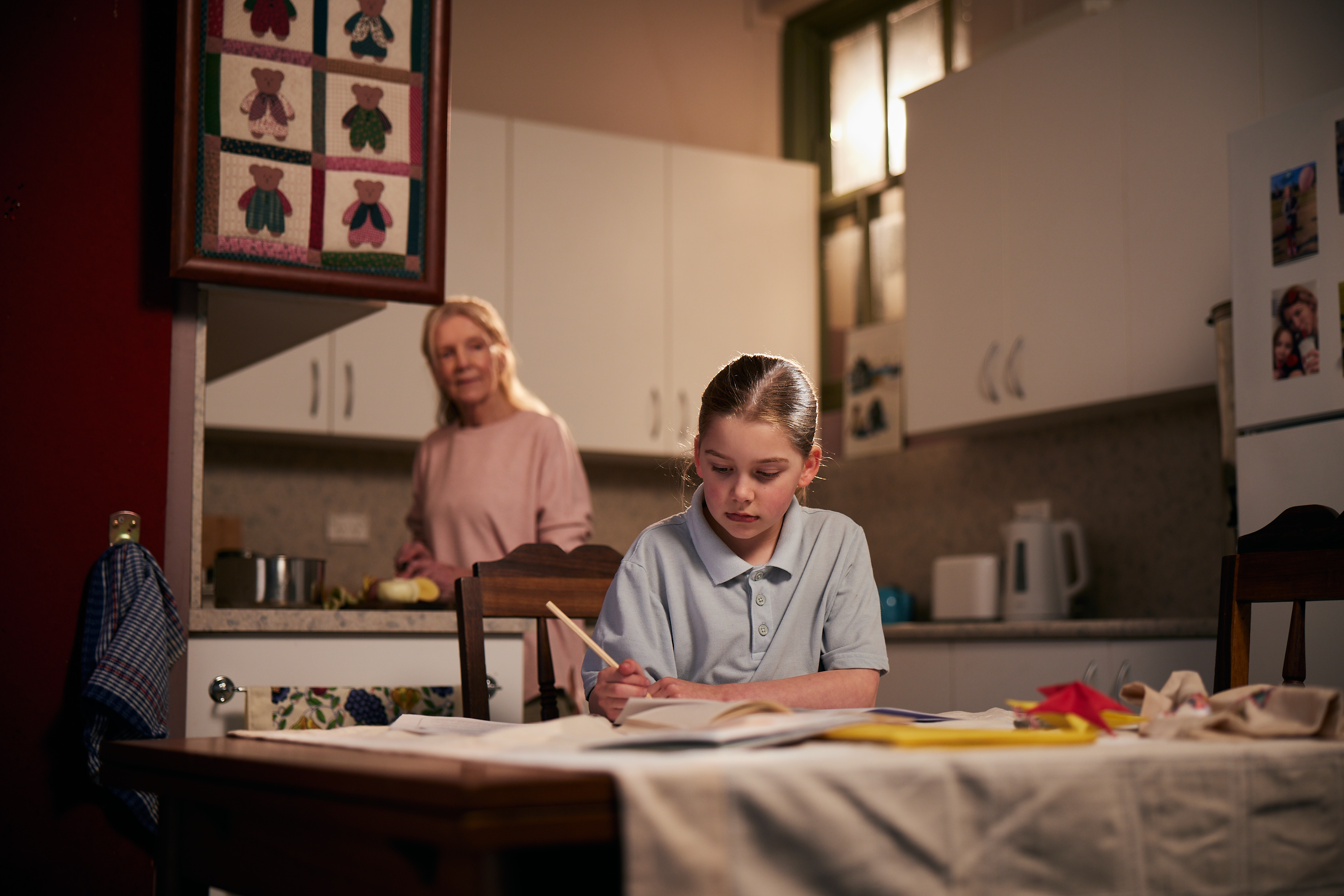 Student doing homework at a table at home with an adult in the background.
