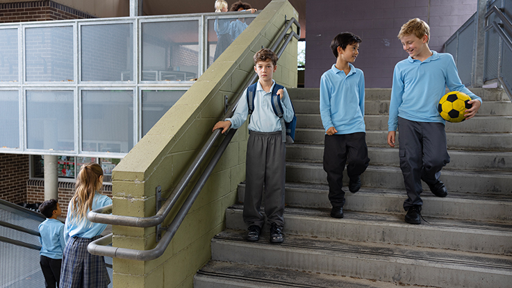 Student standing alone on school stairs while other students walk together nearby.