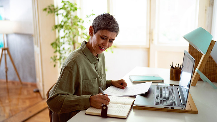 a woman is writing on her notebook in front of her laptop