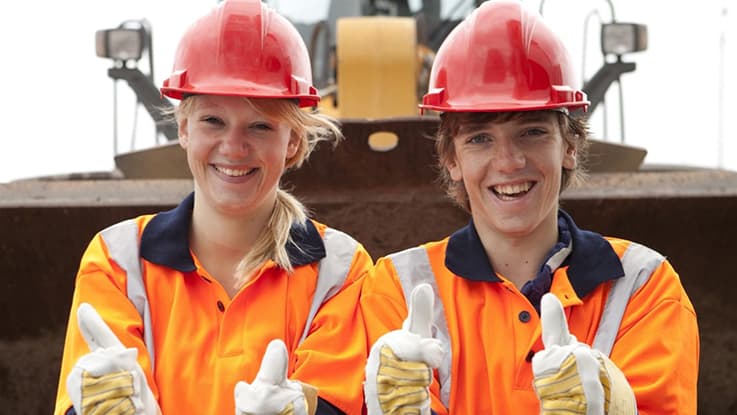 two students with high-vis and red hard hats with gloves and their thumbs up