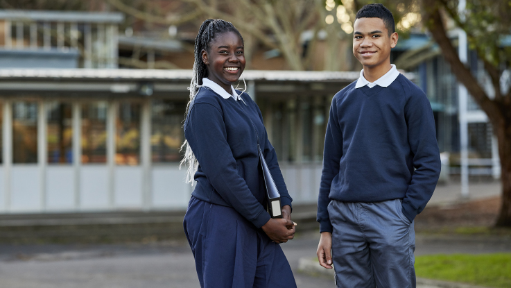 Two students standing in school grounds smiling at camera wearing school uniform