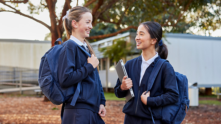 students smiling at each other in school grounds holding school items and wearing backpacks