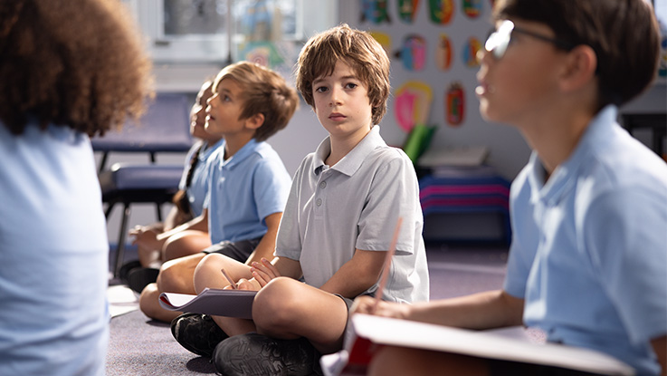 a boy looking neutral sitting on the floor with his peers in the classroom