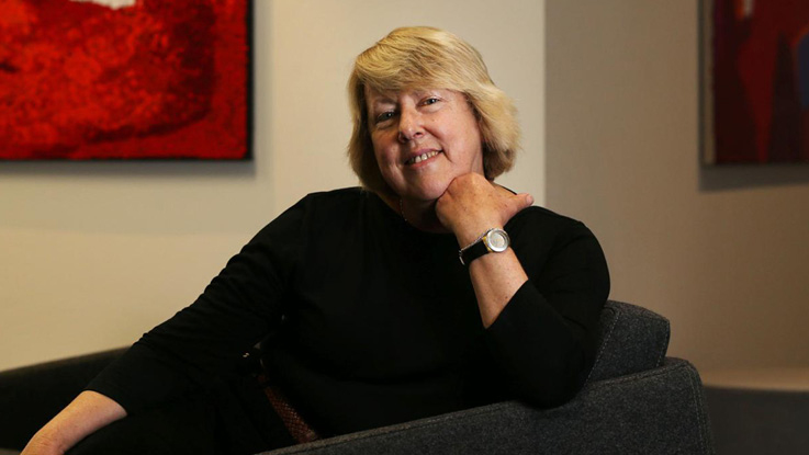 Person seated in an armchair, posing for a portrait in an indoor setting with red artwork on the wall behind.