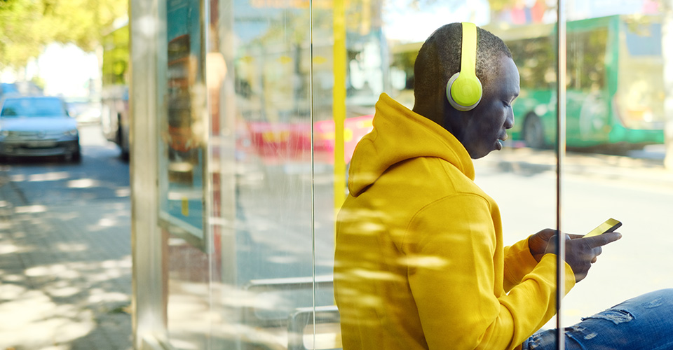 man sitting at a bus stop listening to music with yellow headphones on his phone