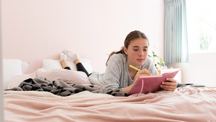 a teenage girl is writing on her notebook while lying comfortably on her bed