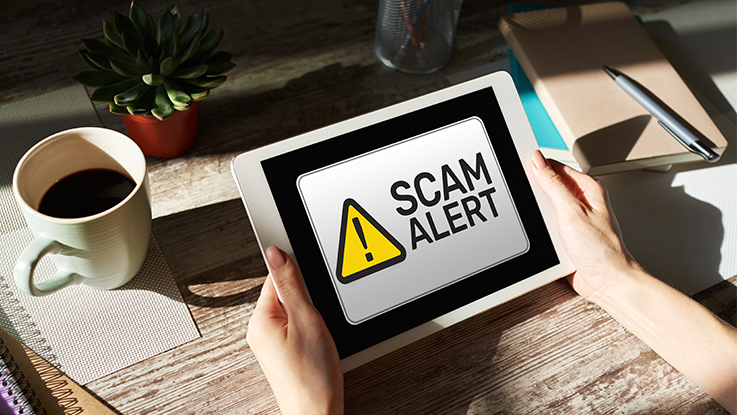 Person holding a tablet displaying a scam alert warning message on a desk with a coffee cup and office items nearby.