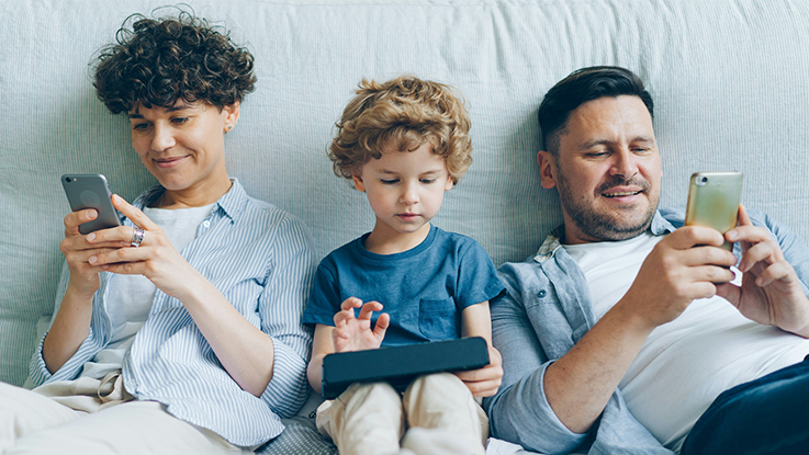 A parent and two kids looking at screens sitting on a bed