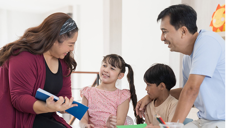 A woman holding a blue book is sitting and talking to two children, while a man stands nearby. The background shows a bright room with some colorful decorations.