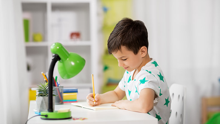 Young child sitting at a desk, writing in a notebook with a pencil under a green desk lamp.