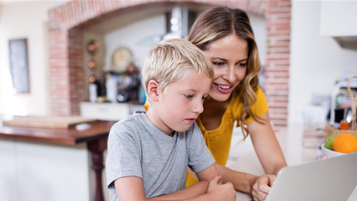Adult sitting with a child using a laptop together at a kitchen table.