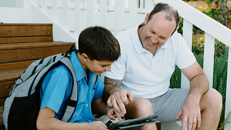 Adult and school-aged boy sitting together on outdoor steps using a tablet, smiling and learning together before school.
