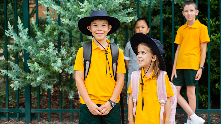 Primary school children in yellow school uniforms and hats standing outside a school fence with backpacks, smiling and talking together.