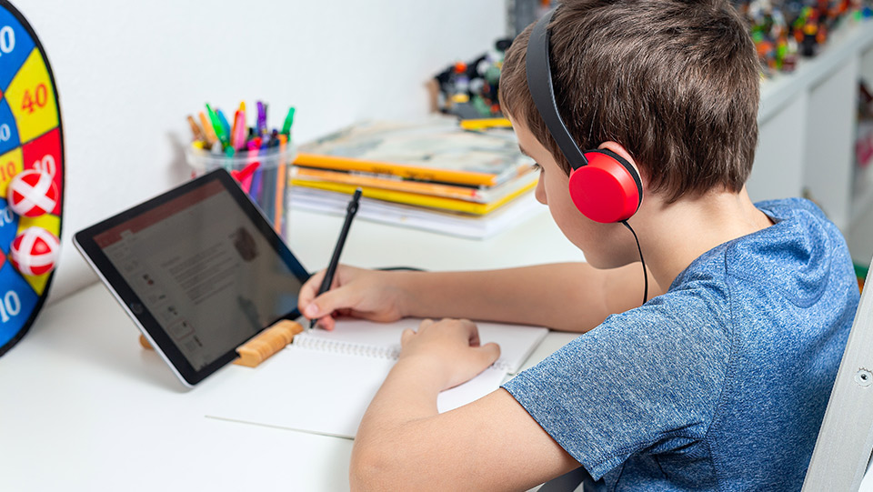 a boy listening to music with over ear headphones doing homework with his tablet on a desk