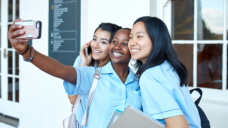 Three schoolgirls in uniform smiling and taking a selfie together outside a school building