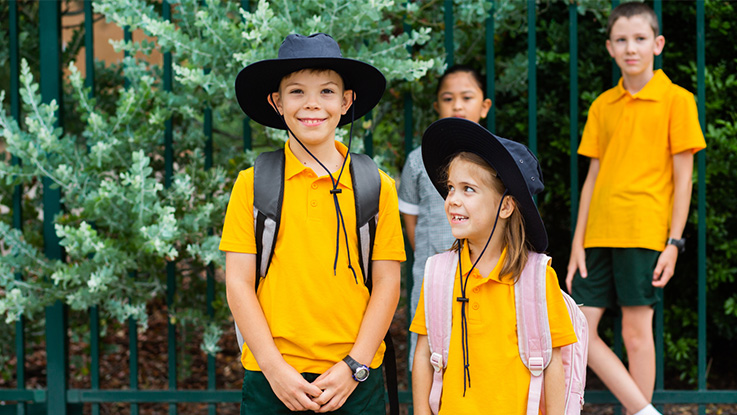 two school children at the front, a boy smiling while the girl is looking up peering at him, two children in the foreground standing next to each other