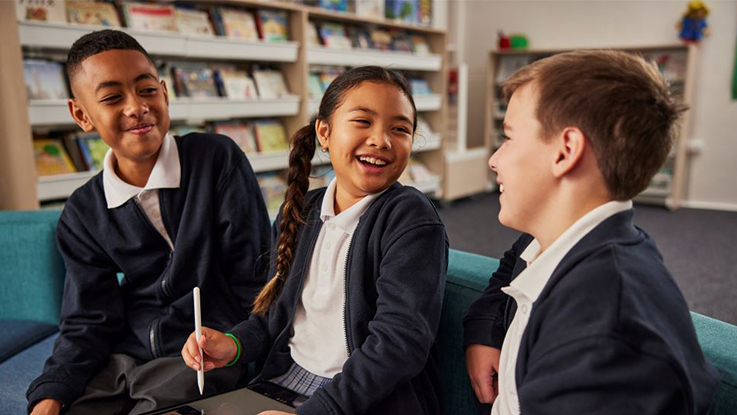 Three children in school uniforms sitting on a couch in a library, with bookshelves in the background. One child is holding a stylus and looking at a tablet.