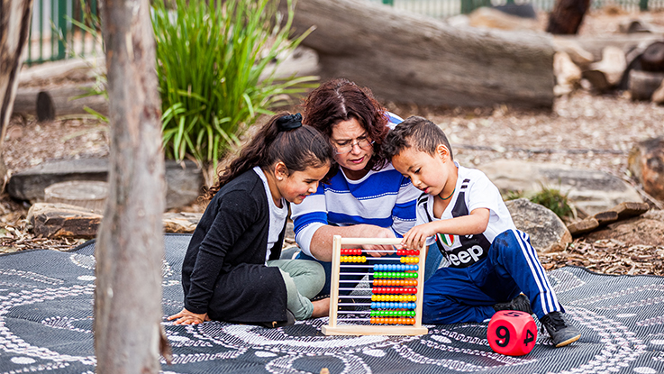 Three children sitting together outdoors, reading a book on a playground surface.