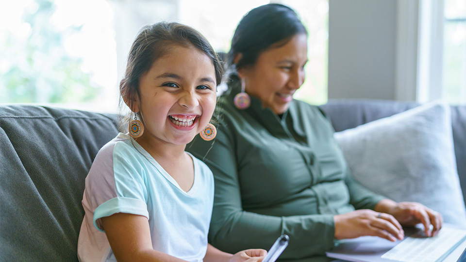 mother and daughter sitting on the couch together. the mother is sitting on the laptop and the daughter is drawing next to her looking directly at the camera
