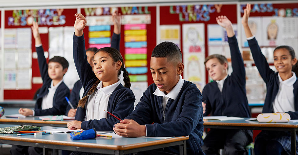 a group of school students with their hands up with the student at the front heads down doing work
