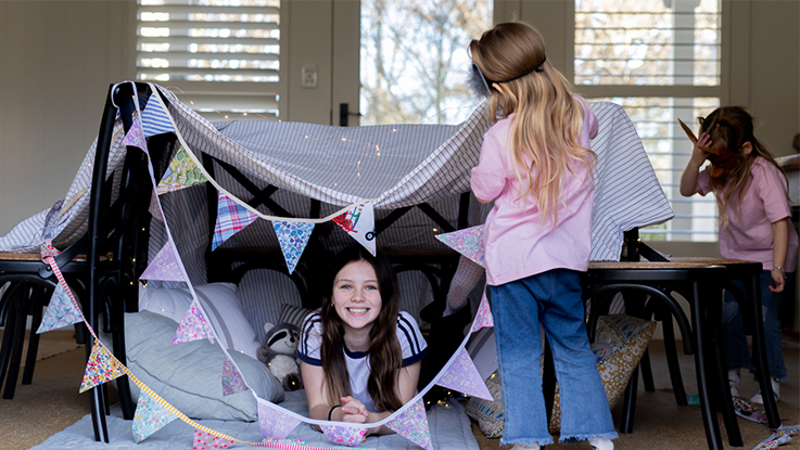 Two children building and playing in a blanket fort indoors, decorated with bunting and cushions.