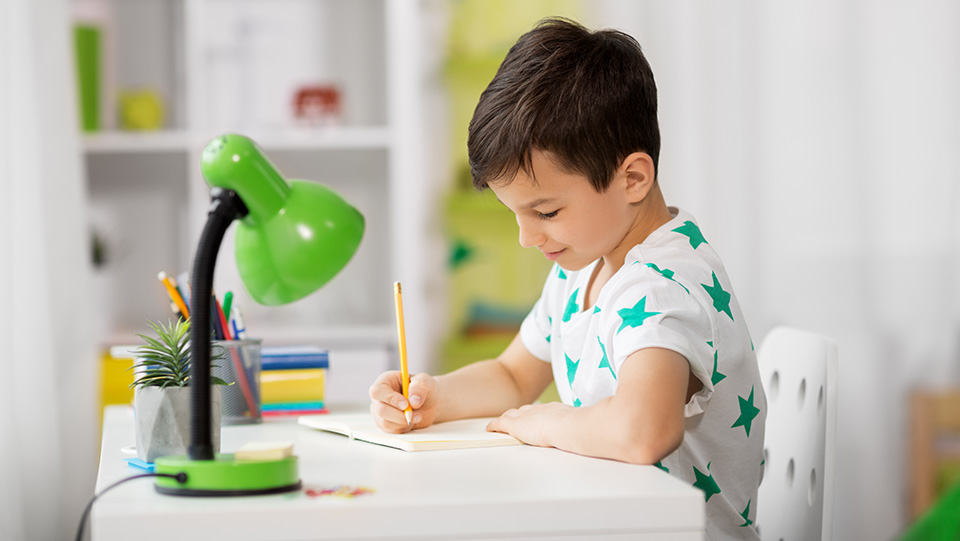 a child is writing in his notebook on a desk that has a green lamp 