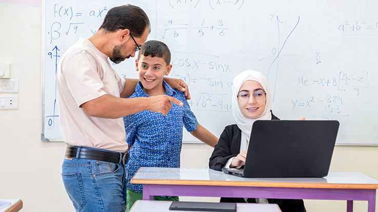 a boy being held by his father who is pointing at the teacher laptop in a classroom