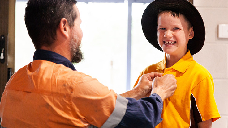 a father who is wearing high vis is helping his young son button his school yellow polo