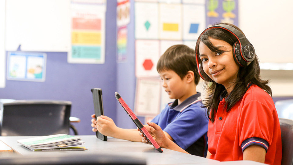 a school girl wearing a red polo is listening to a audio on her tablet and a school boy is sitting next to her listening on his tablet