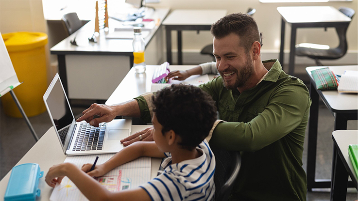 A teacher assists a student with a laptop in a classroom setting. The student is writing in a notebook while the teacher points at the laptop screen. The classroom has desks, chairs, and various school supplies visible.