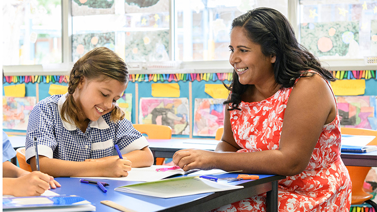 Child and adult seated together at a table in a classroom, looking at educational materials, with colourful student artwork displayed on the wall in the background.
