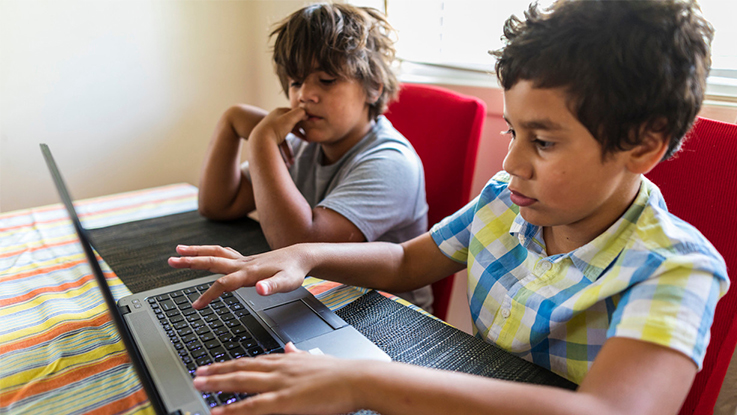 Two children using a laptop together at a table, with one child pointing at the screen while the other uses the keyboard. Colourful books or learning materials are stacked nearby indoors.