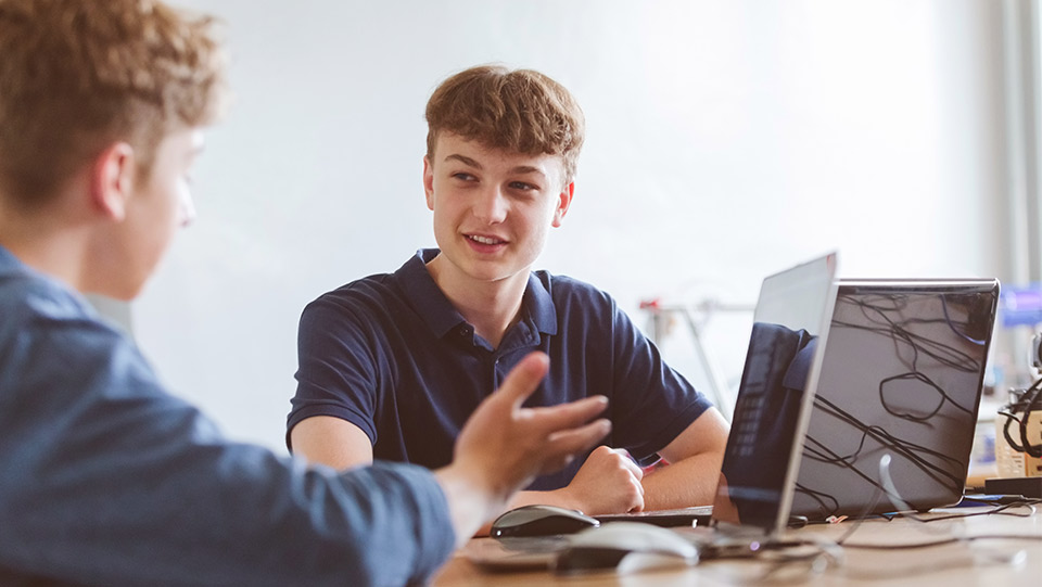 two male teenagers who are chatting at a desk with their laptops