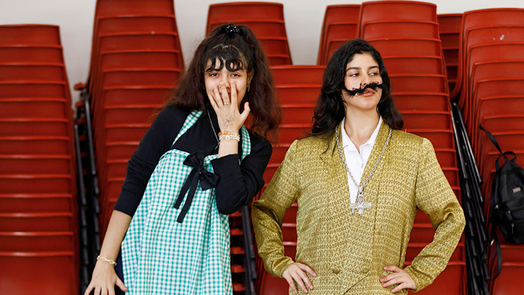 Two people in theatrical costumes pose in front of stacks of red chairs.