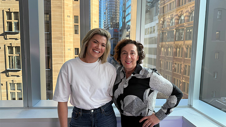 two women posing together. the woman on the left has short blonde hair and is wearing a white t shirt with jeans and the woman on the right has brown medium curly hair wearing a black and white blouse with dark pants