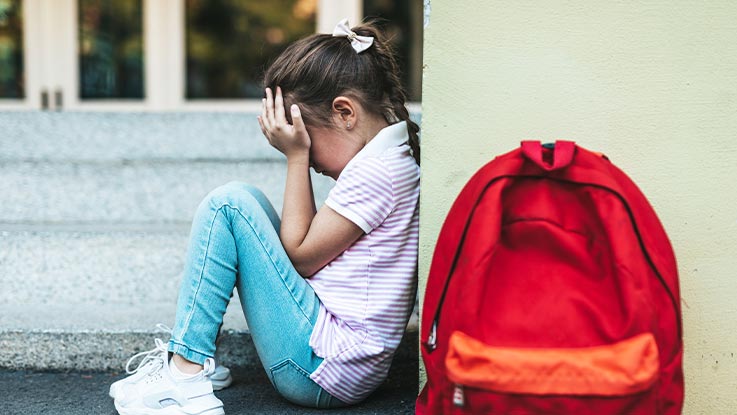 Sad small girl seating on floor with hand covering face and next to her there is read backpack