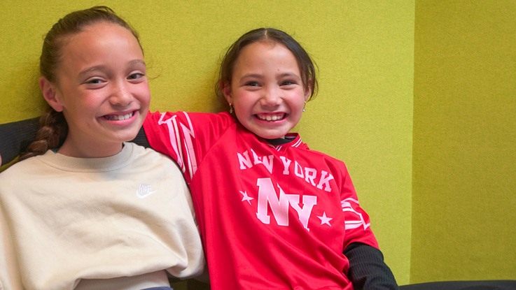 Two children sit together smiling against a green wall.
