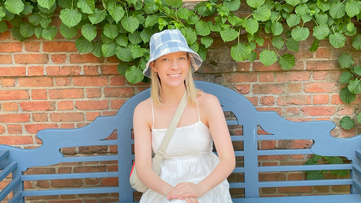 a young woman sitting on a blue bench outside wearing a blue bucket hat, white dress and white crossover bag