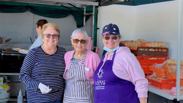 three ladies posing together at a bbq, the woman on the far left is wearing a blue striped top, the lady in the middle is wearing a pink cardigan with a stripped top and the far right lady is wearing a view apron, with a pink jumper