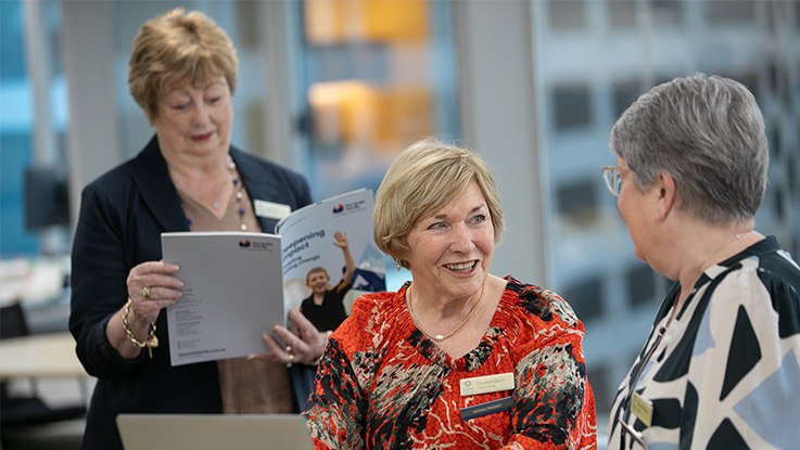 a woman in a red top on a laptop talking to her colleague while their colleague is in the background reading