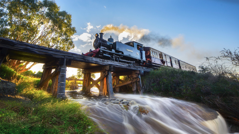 Historic steam train crossing a timber bridge over a rushing creek, surrounded by trees and countryside.